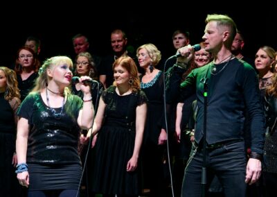 Duettopari laulaa mikrofoneihin toisiaan katsoen. Kuoro taustalla.