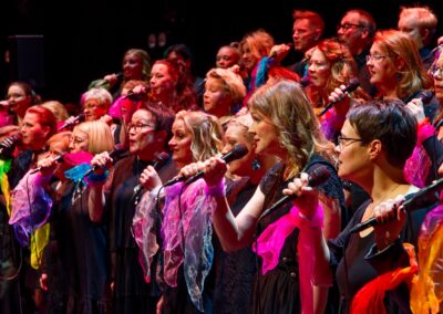Kuorolaiset laulamassa mikrofoneihin. Mustiin pukeutuneilla kuorolaisilla värikkäät huivit ranteeseen solmittuna.