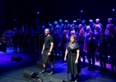 Duettopari laulaa mikrofoneihin lavalla sinisissä valoissa. Taustalla kuoro.